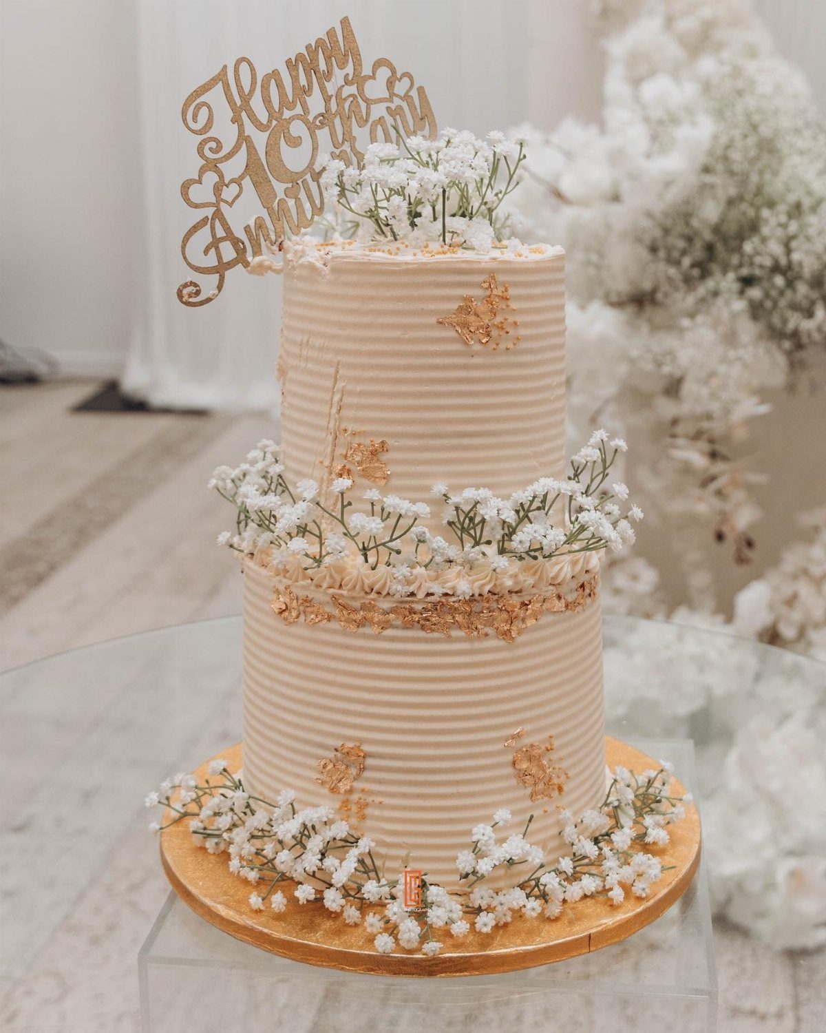 Two-tier cream anniversary cake with gold leaf, white flowers, and 10th anniversary topper.