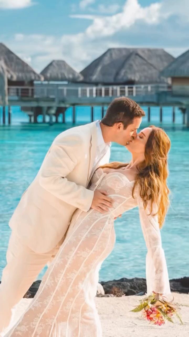Couple in white wedding attire kissing on a beach with overwater bungalows.