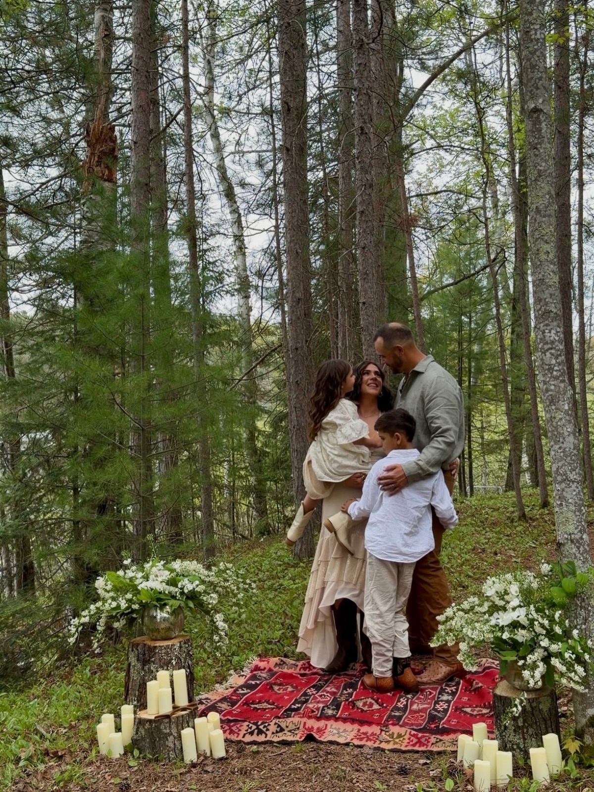 Family embracing on a red rug in a forest with candles and flowers