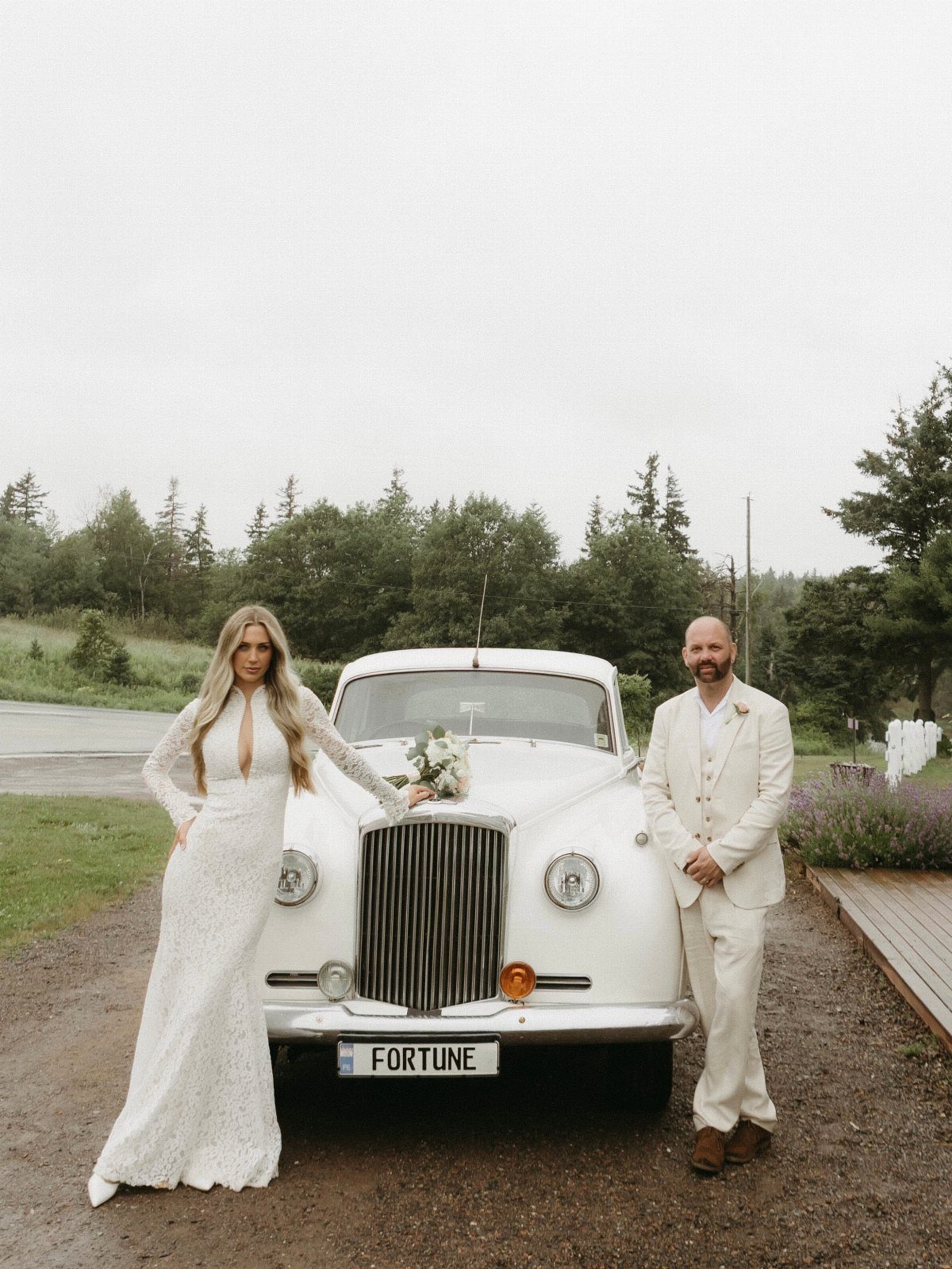 Couple in wedding attire posing in front of a vintage white car outdoors.
