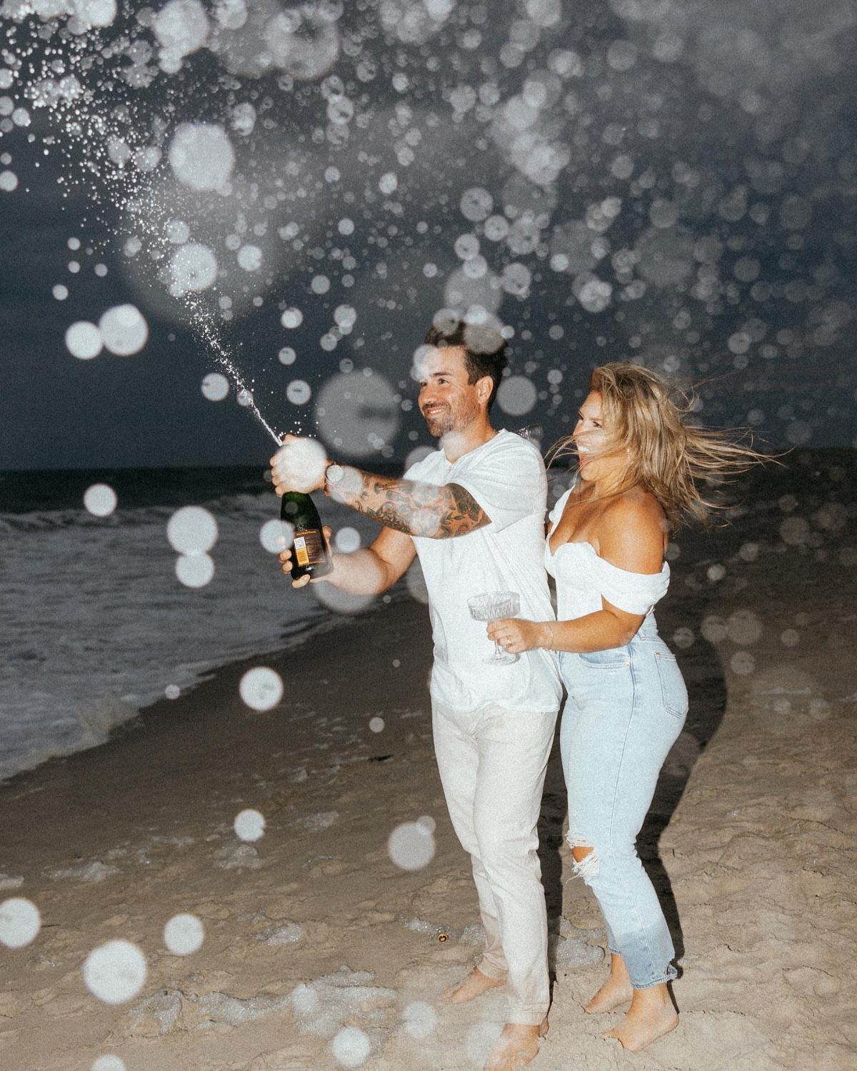 Couple celebrating on a beach at night with a popping champagne bottle spray