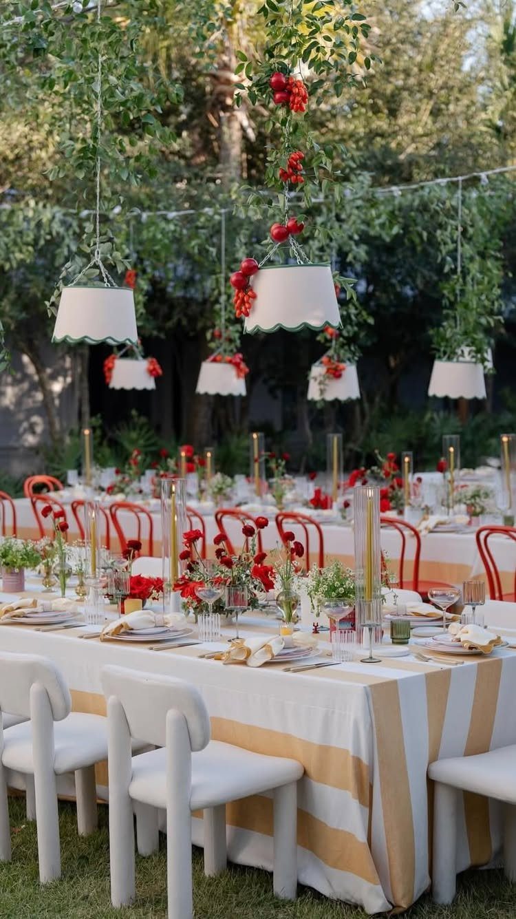 Outdoor dining tables with striped linens, red flowers, and hanging lamps in a garden.