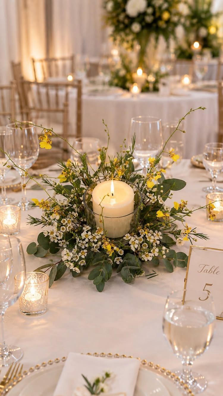 Floral centerpiece with white and yellow flowers and a lit candle on a table.