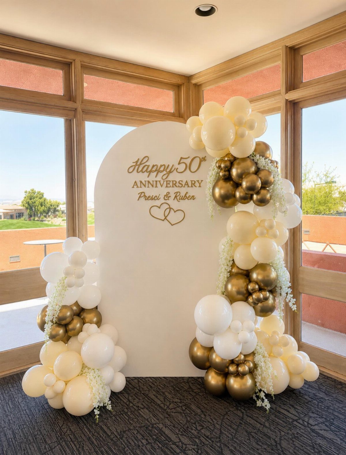 White arched anniversary backdrop decorated with gold and white balloons and floral accents.