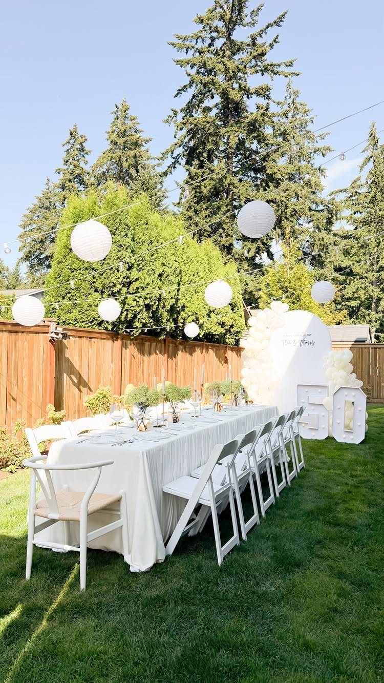 Outdoor party table with white linens, gold 50 numbers, and hanging white lanterns.