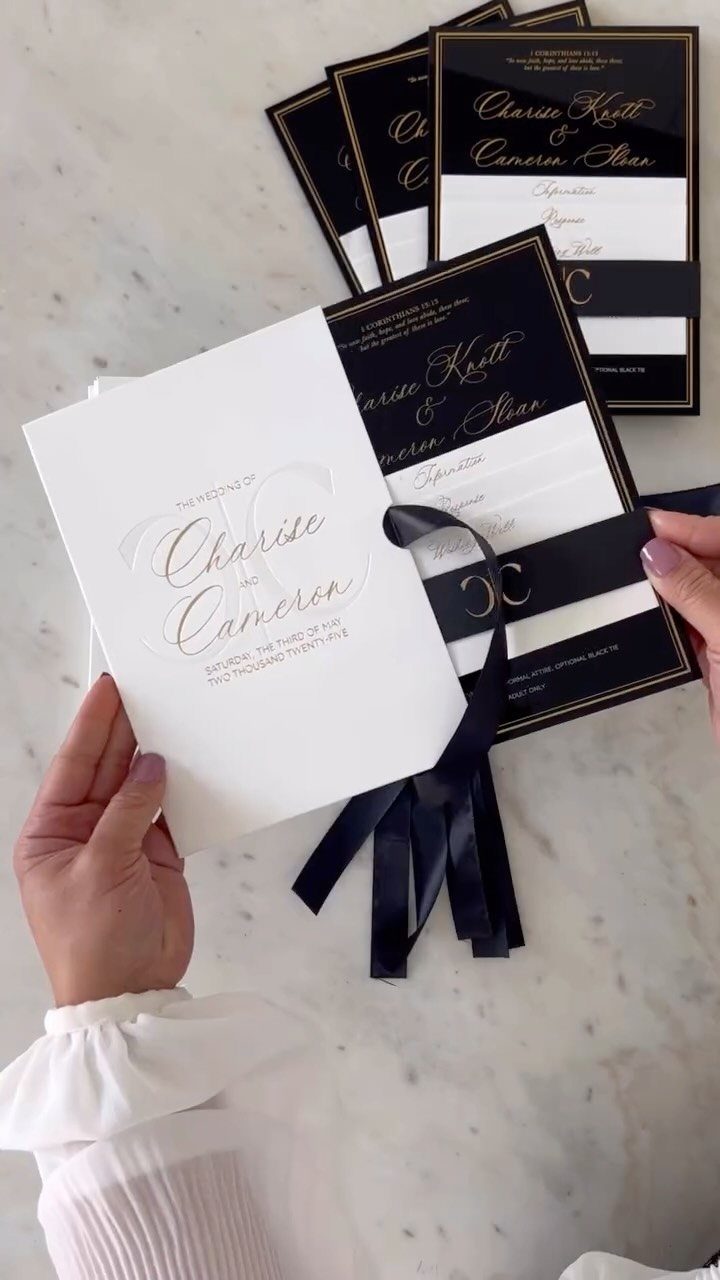 Wedding invitations with gold script and black ribbons on a white marble surface.