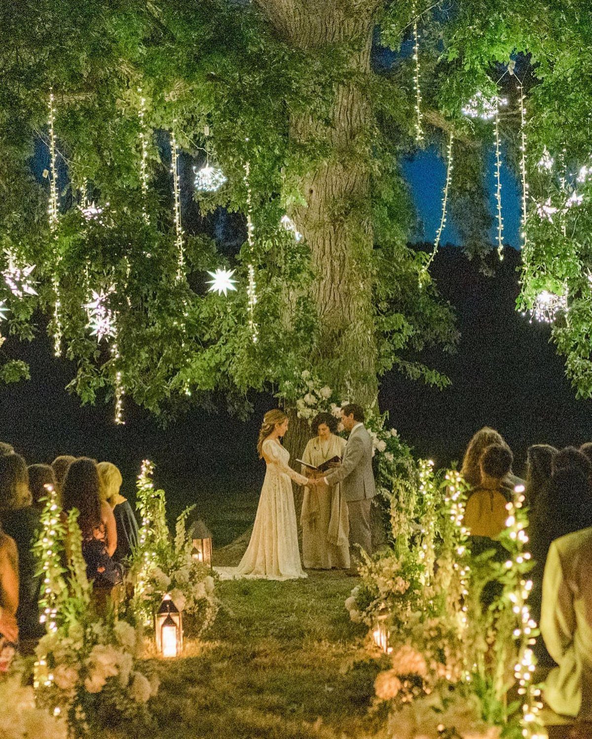 Couple exchanging vows under a large tree decorated with fairy lights and flowers.
