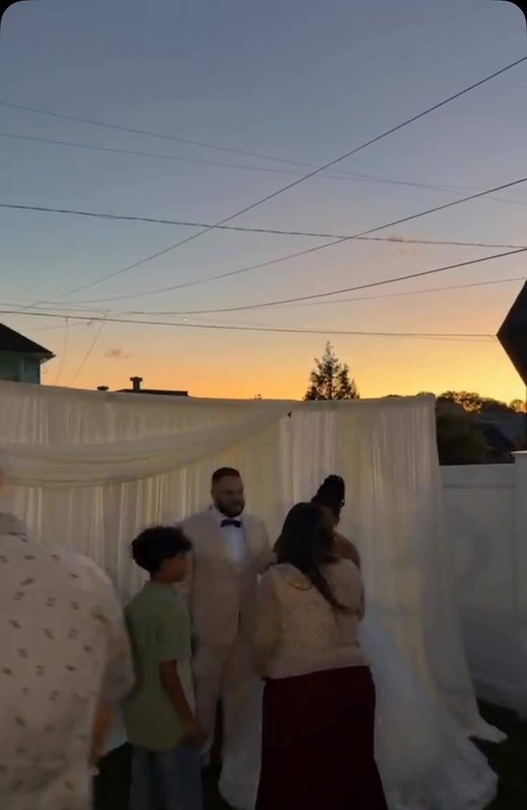 Couple exchanging vows in a backyard with a white backdrop during a golden sunset.