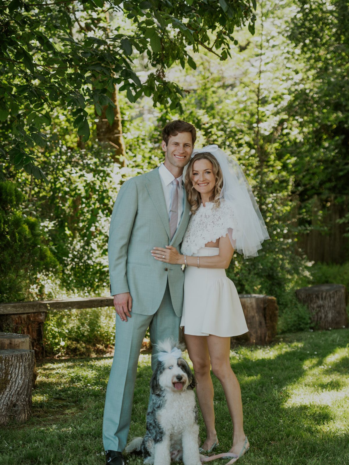 Couple and dog posing in a garden, woman wearing a short white wedding dress.