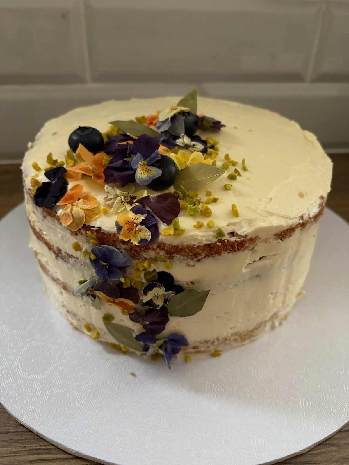 Semi-naked layer cake decorated with blueberries, edible flowers, and pistachio crumbles.