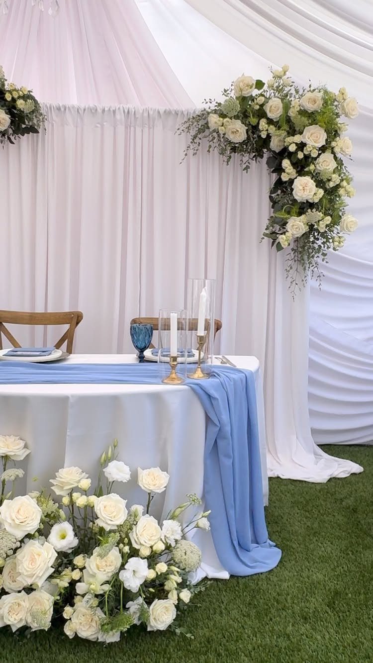 Small white wedding table for two with blue runner and white floral arrangements.
