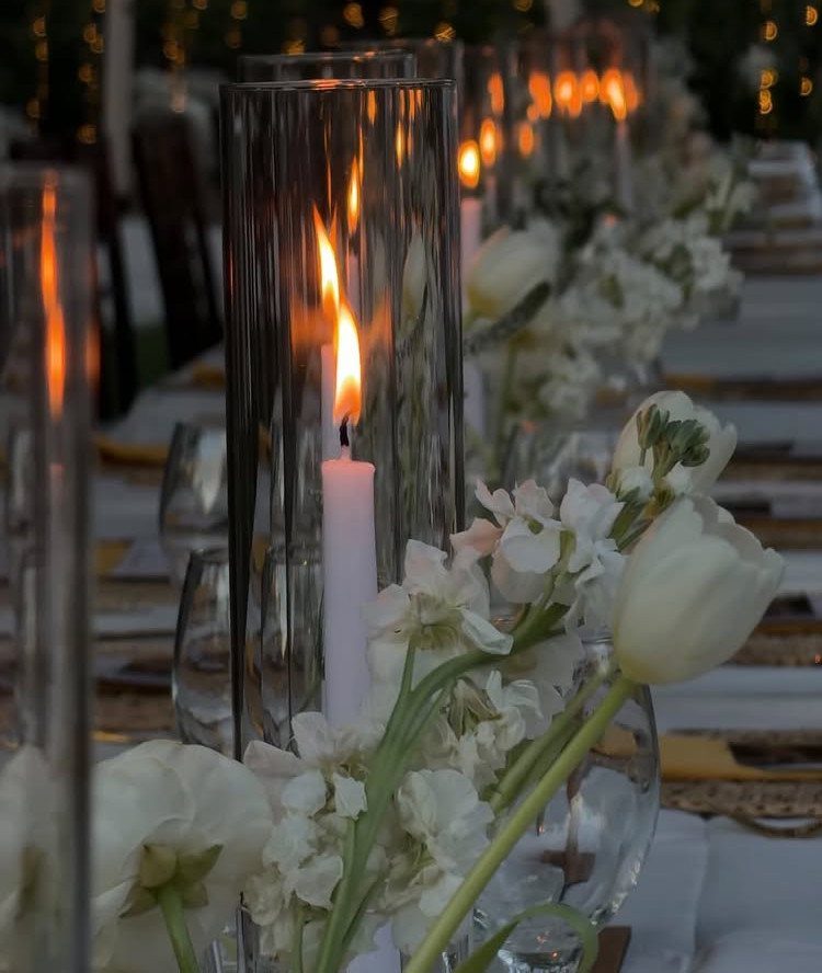 Lit white taper candles in glass cylinders with white flowers on a table