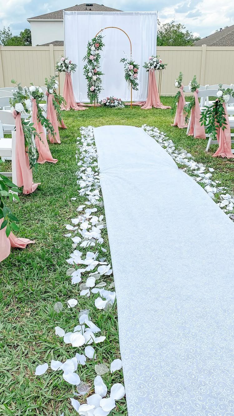 Wedding ceremony aisle with white petals, pink chair decor, and floral gold arch.