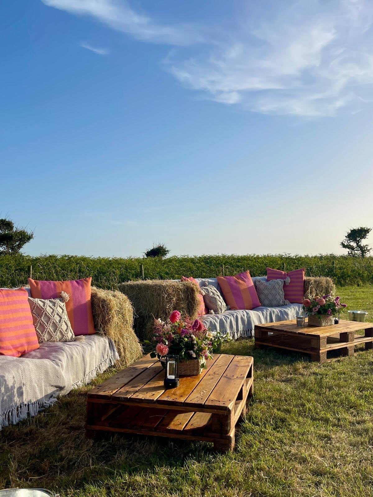 Hay bale seating with pink pillows and wooden pallet tables in a field