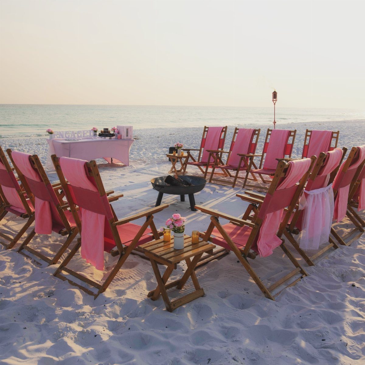 Wooden beach chairs with pink fabric surrounding a black fire pit on white sand.