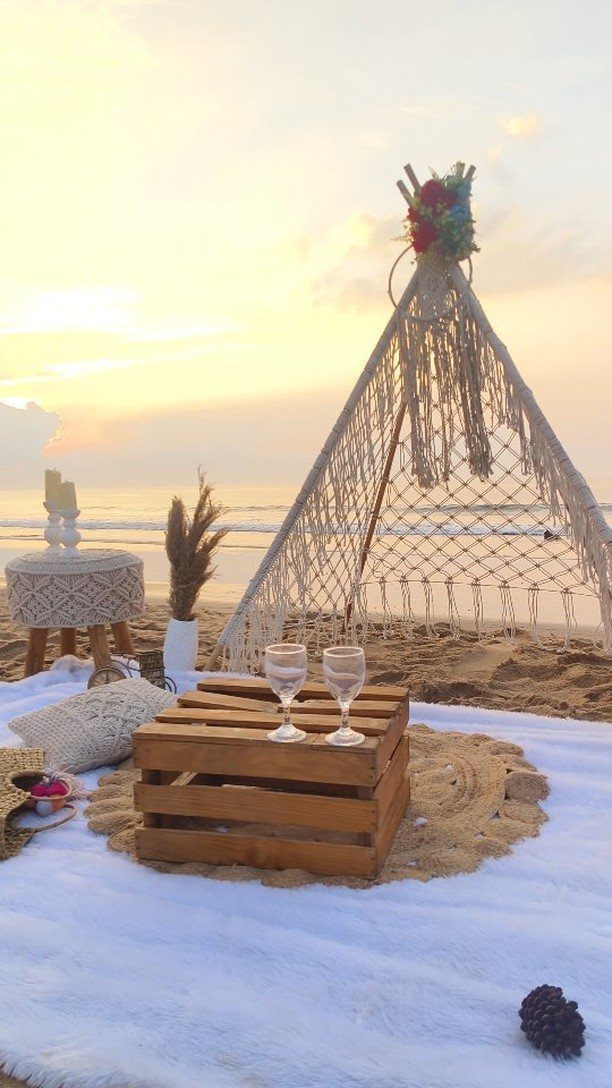 Beach setup with a macrame teepee, wooden crate table, and wine glasses at sunset.