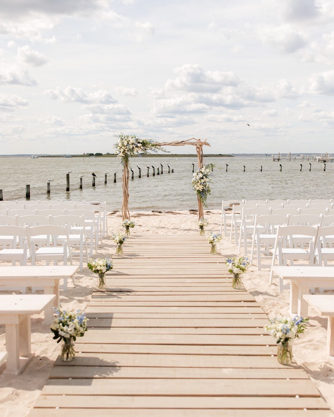 Driftwood arch with white flowers at the end of a wooden beach aisle.