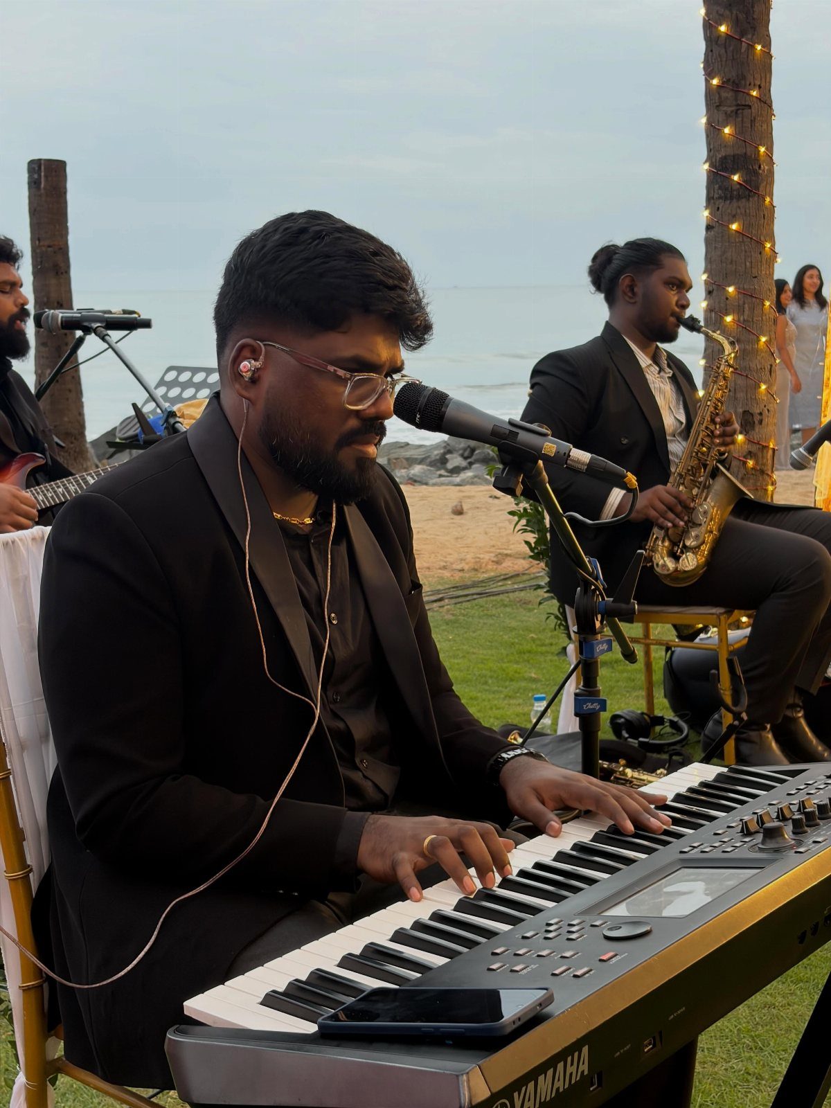 Musicians in black suits playing keyboard and saxophone at an outdoor beach event.
