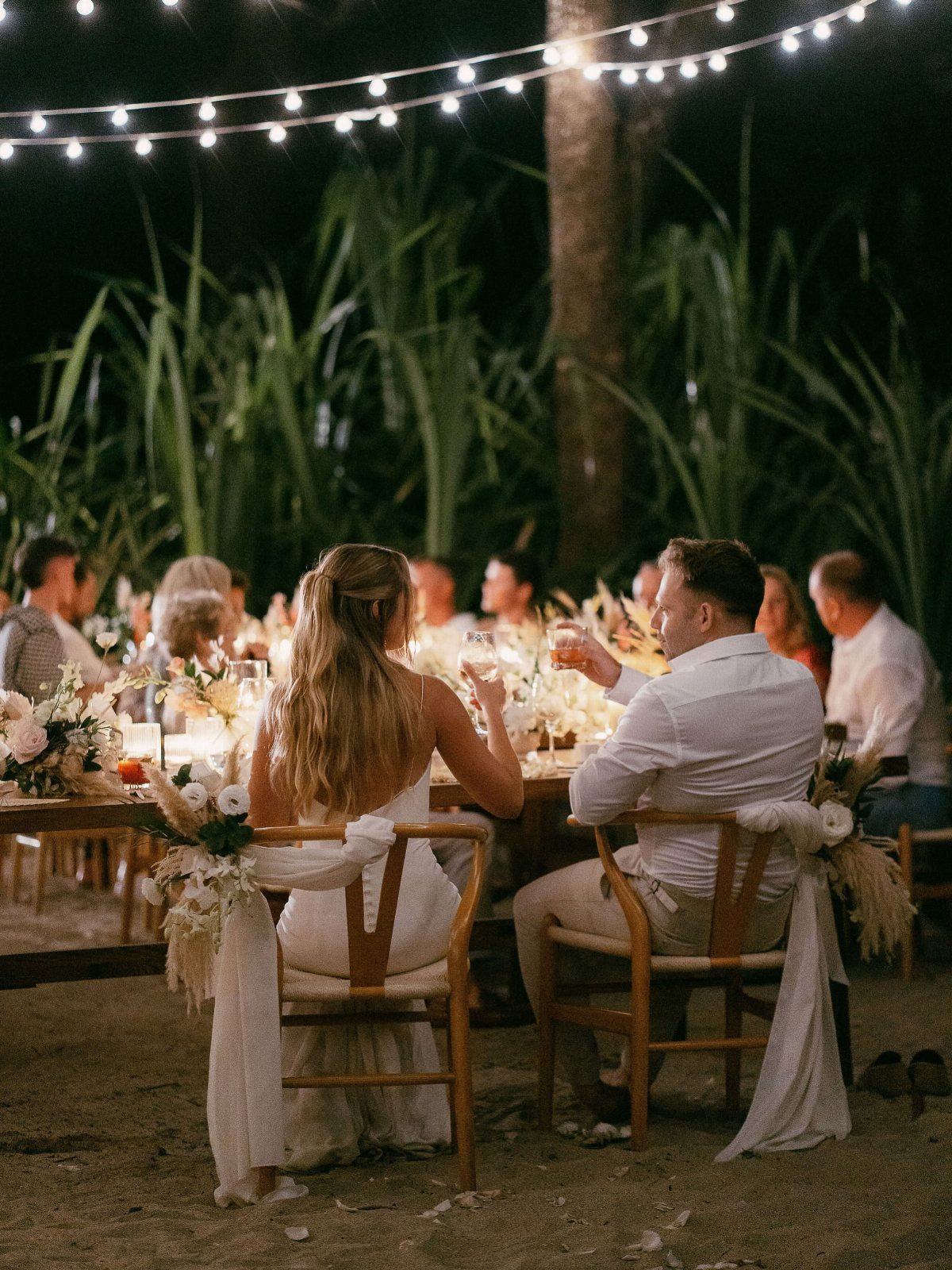 Wedding couple dining at a beach table under strings of warm fairy lights.