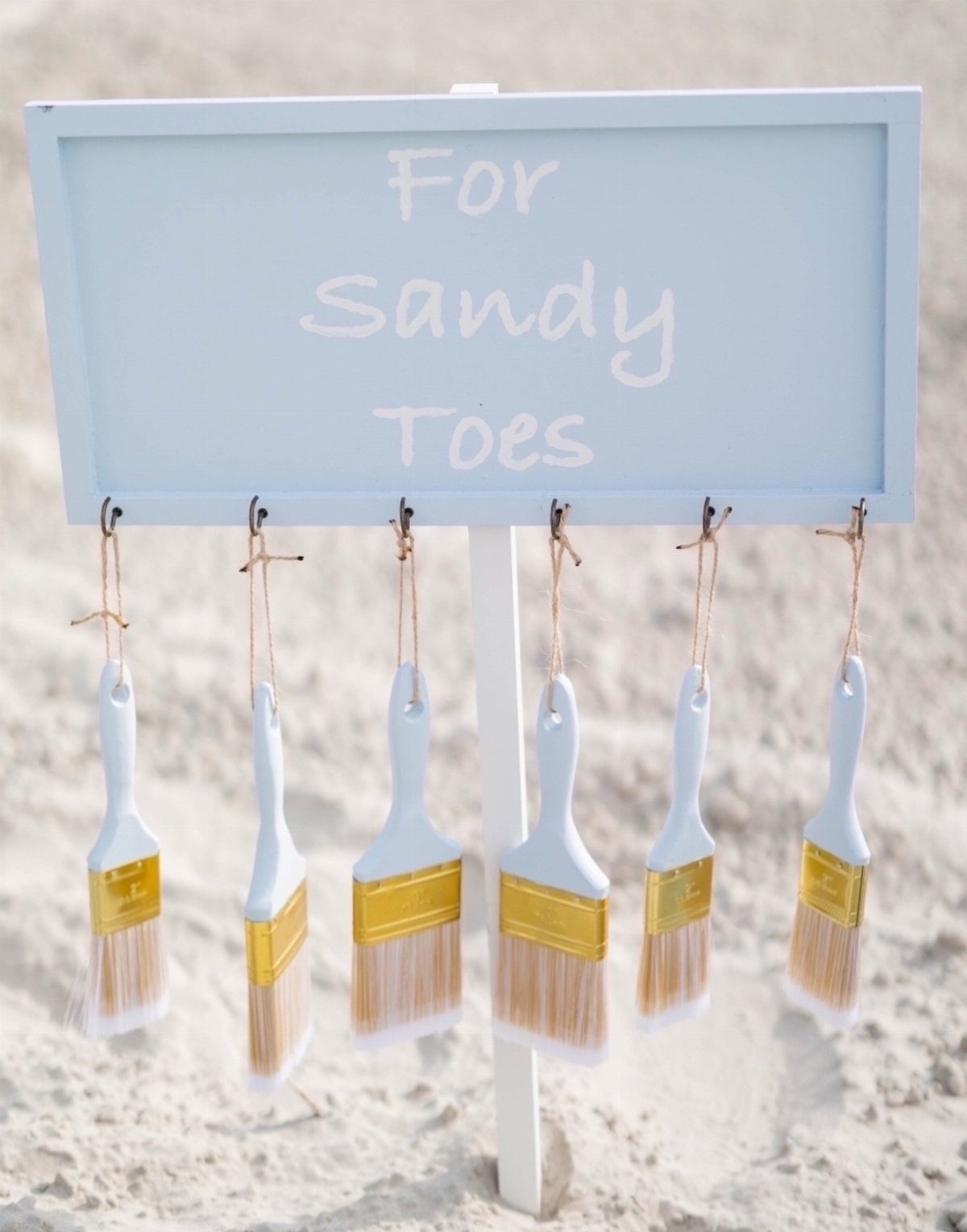 Light blue sign with paintbrushes hanging below it on a sandy beach.