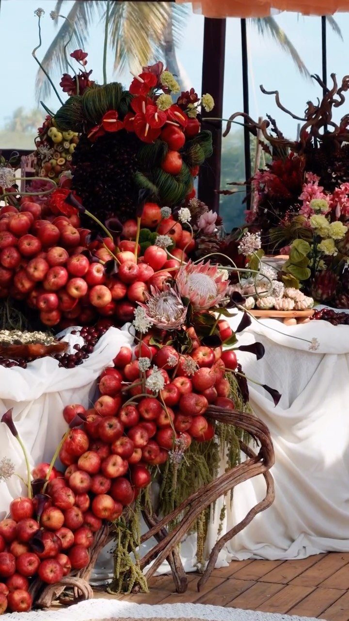 Red apple and tropical flower centerpieces on a white draped table outdoors.