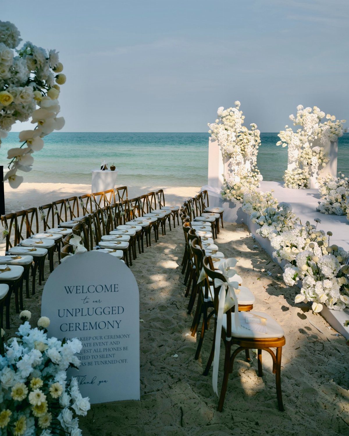 Wooden chairs and white floral arrangements for a beach wedding ceremony by the ocean.