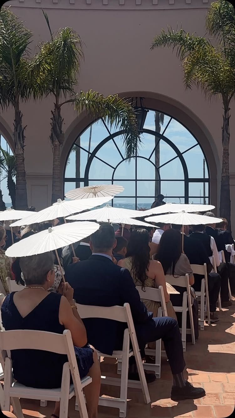 Wedding guests sitting in white chairs holding white parasols for sun protection.