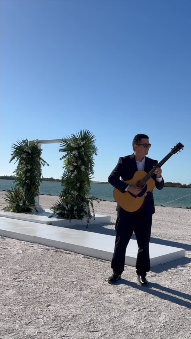 Musician in a suit playing guitar next to a floral wedding arch on a beach