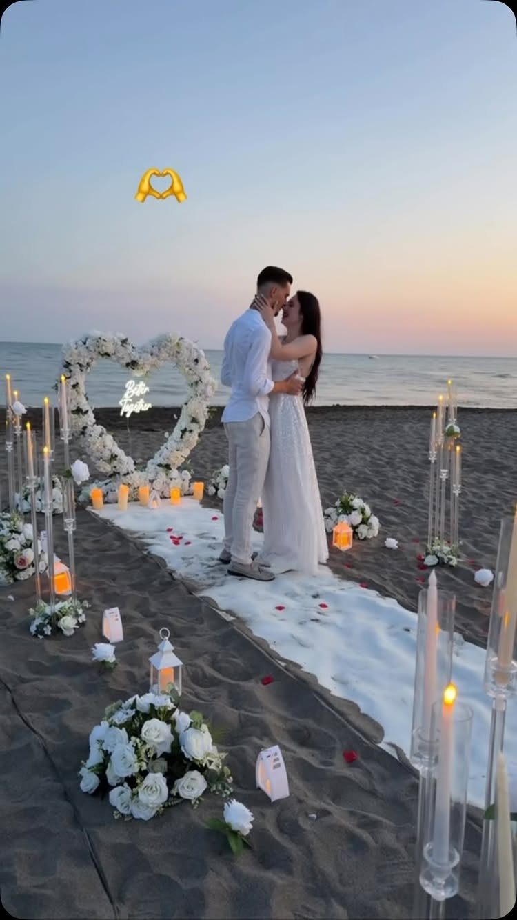 Couple embracing on a beach with a floral heart arch and lit candles.