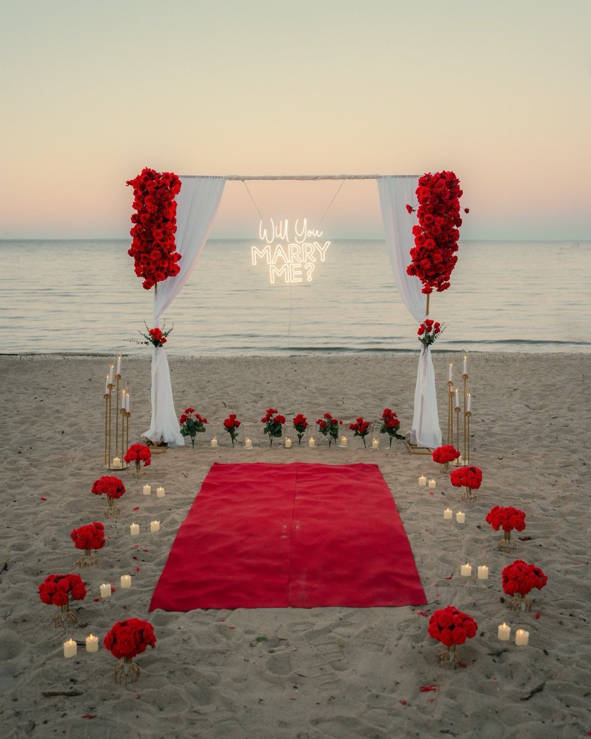 Romantic beach proposal setup with red carpet, roses, and neon marriage sign.