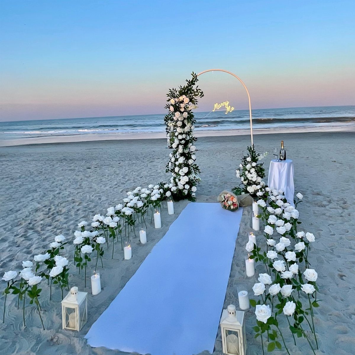 Romantic beach proposal setup with white roses, lanterns, and a floral arch at sunset.