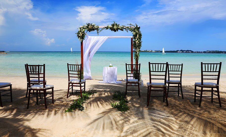 Wedding arch with white flowers and chairs on a sandy beach by turquoise water