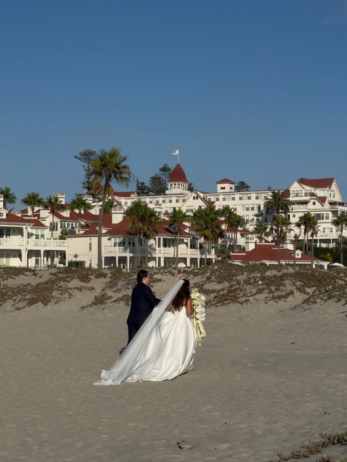 Bride and groom on a sandy beach facing the Hotel del Coronado resort.
