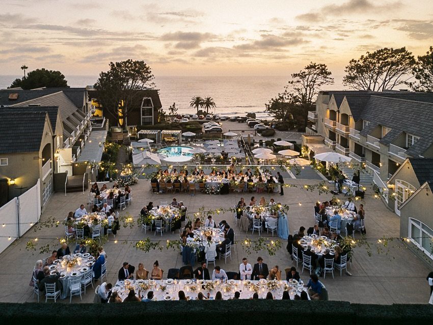 Outdoor wedding reception with guests at decorated tables overlooking the Pacific Ocean at sunset.