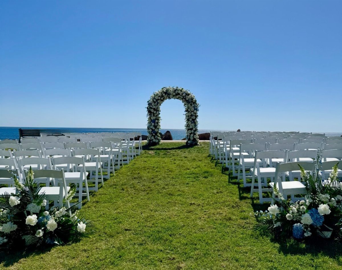 Outdoor wedding ceremony setup with white chairs and a circular floral arch by the ocean