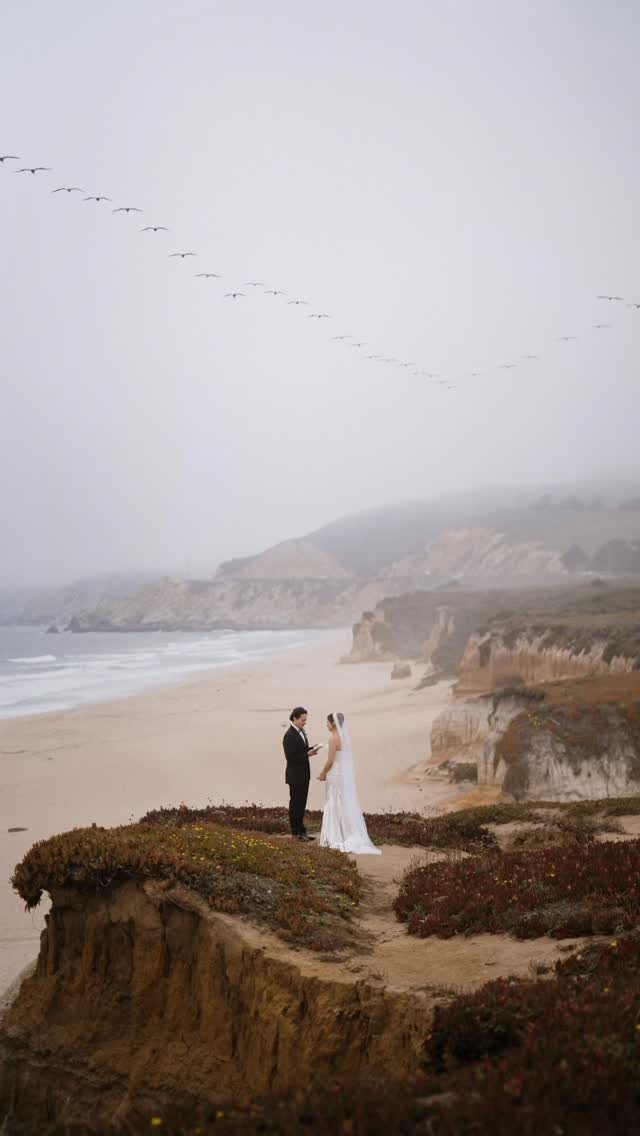 Bride and groom standing on a sandy cliff overlooking a foggy beach coastline