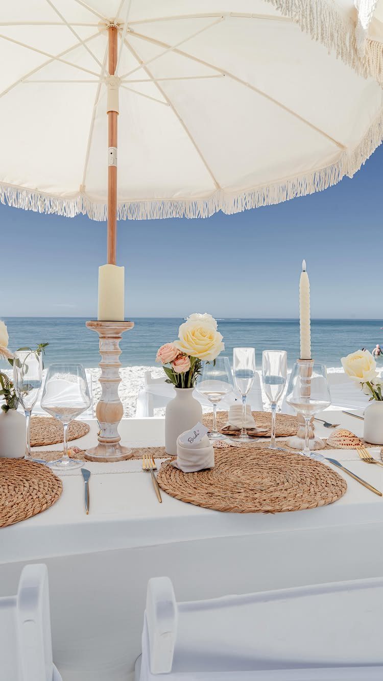 Wedding table setting with white roses and woven placemats under a beach umbrella.