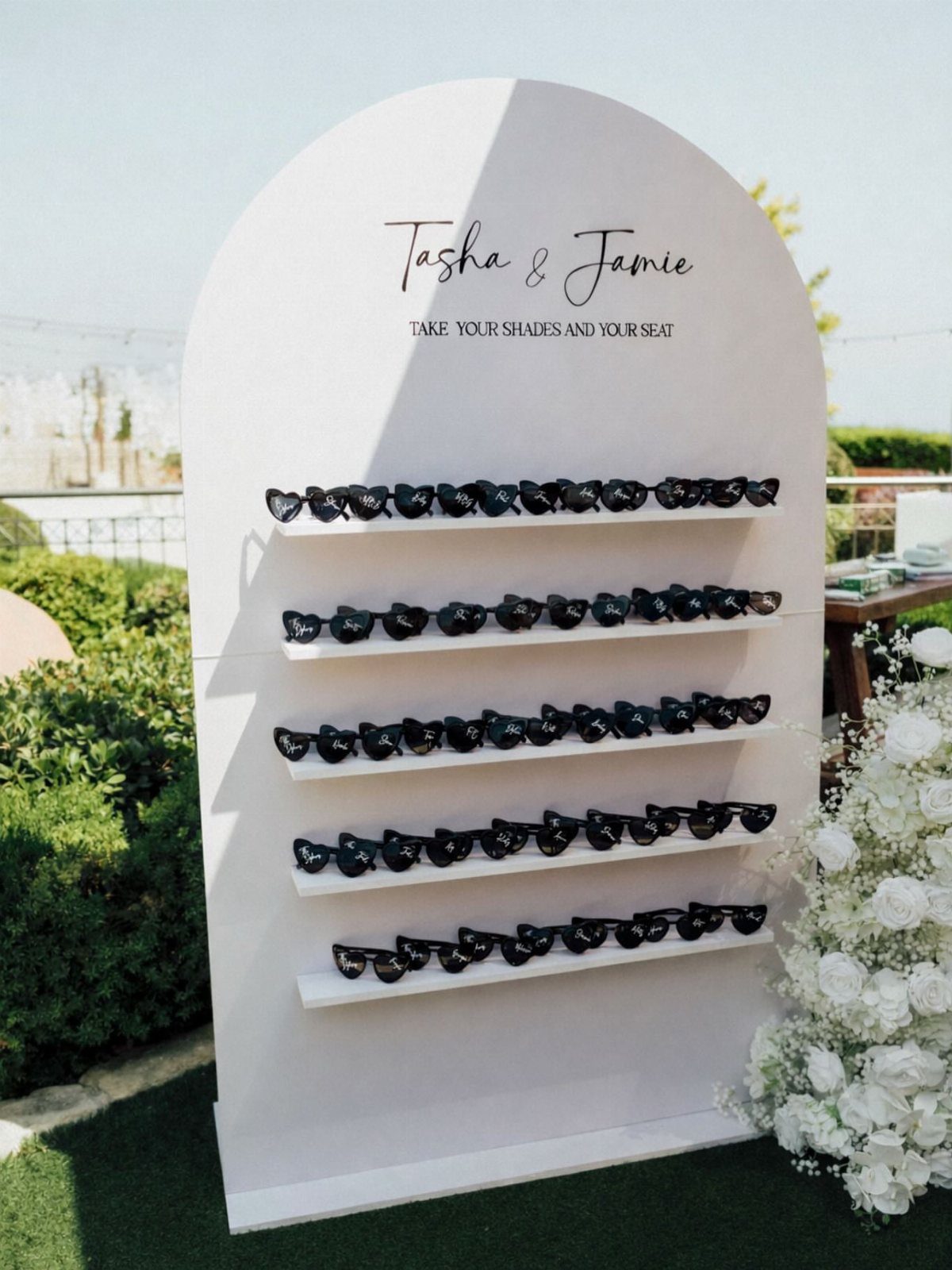 White arched display stand with rows of black sunglasses for a wedding guest station.