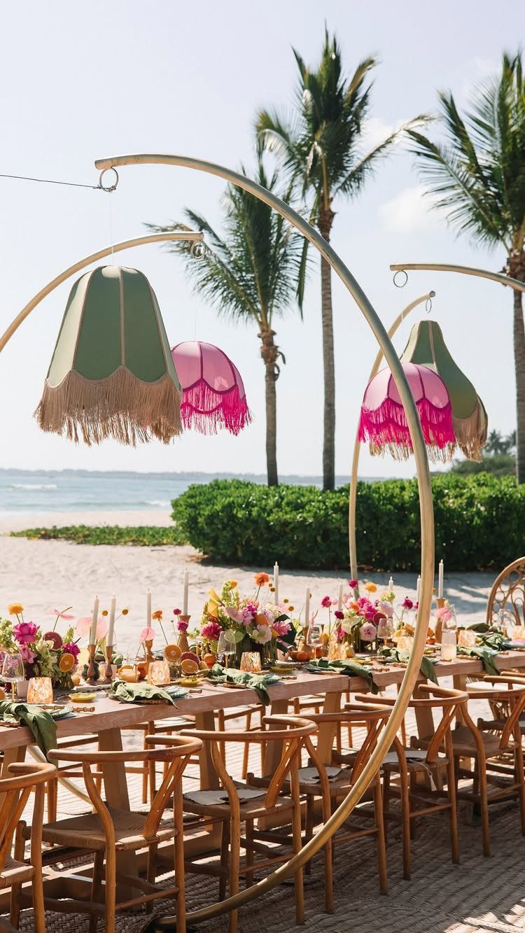 Wedding dining table on a beach with floral centerpieces and hanging fringed lampshades.