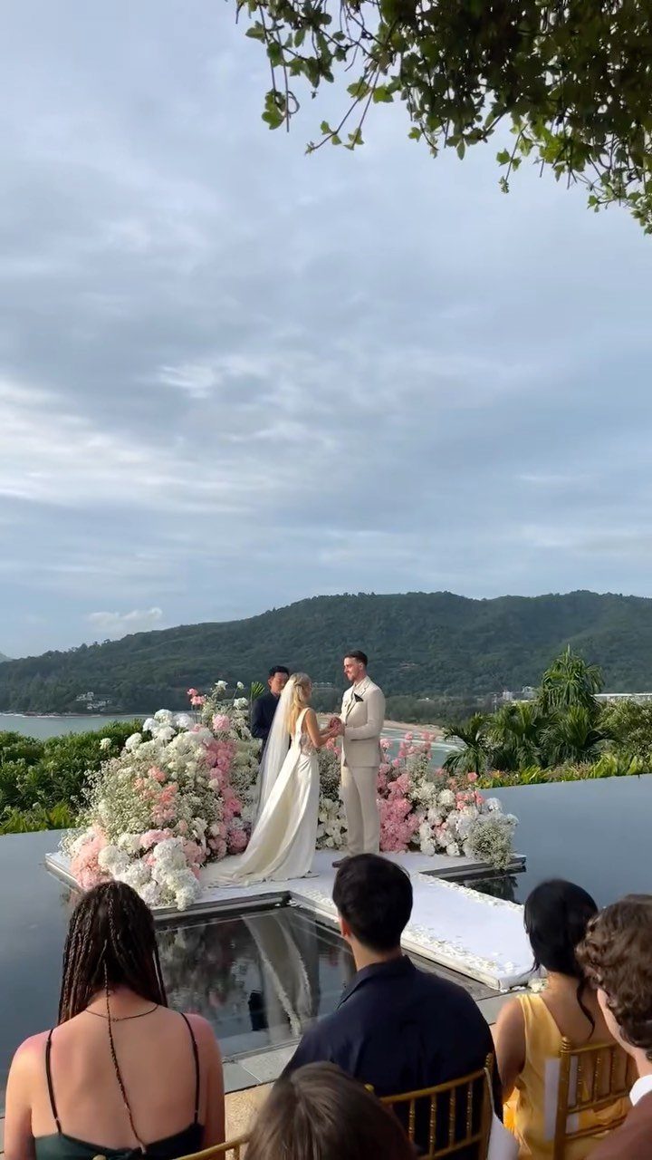 Bride and groom holding hands at a floral wedding altar overlooking coastal mountains.
