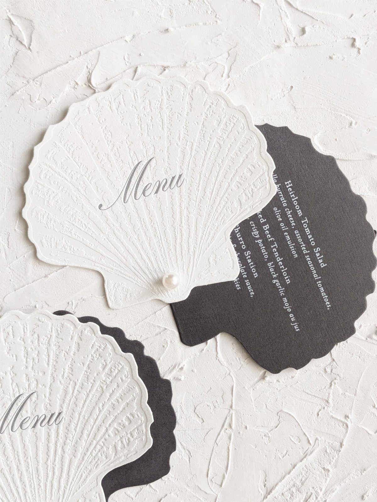 Shell-shaped wedding menu cards in white and black on a textured white background.