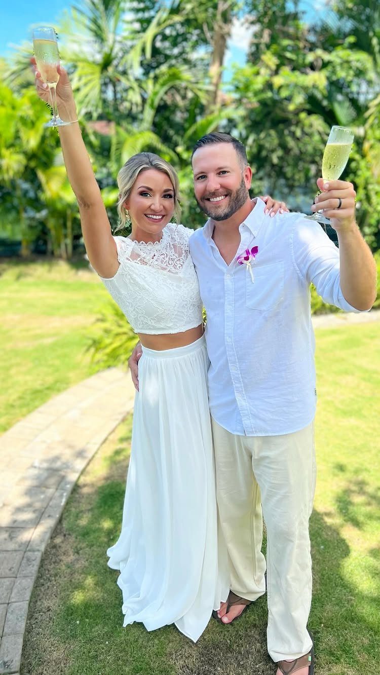 Couple in white beach wedding attire holding champagne glasses in a tropical garden