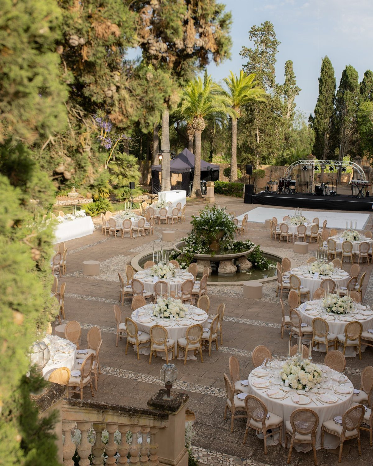 Outdoor courtyard wedding reception with round tables, white florals, and a stone fountain.