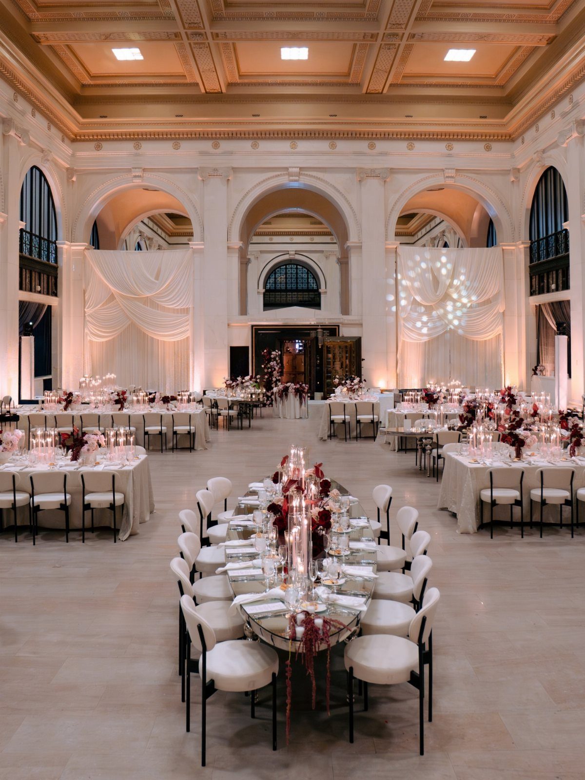 Grand ballroom with white arches, draped fabric, and tables set with red flowers.