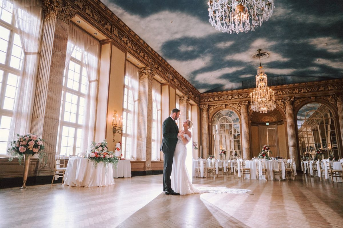 Bride and groom standing in a grand ballroom with cloud ceiling and chandeliers.
