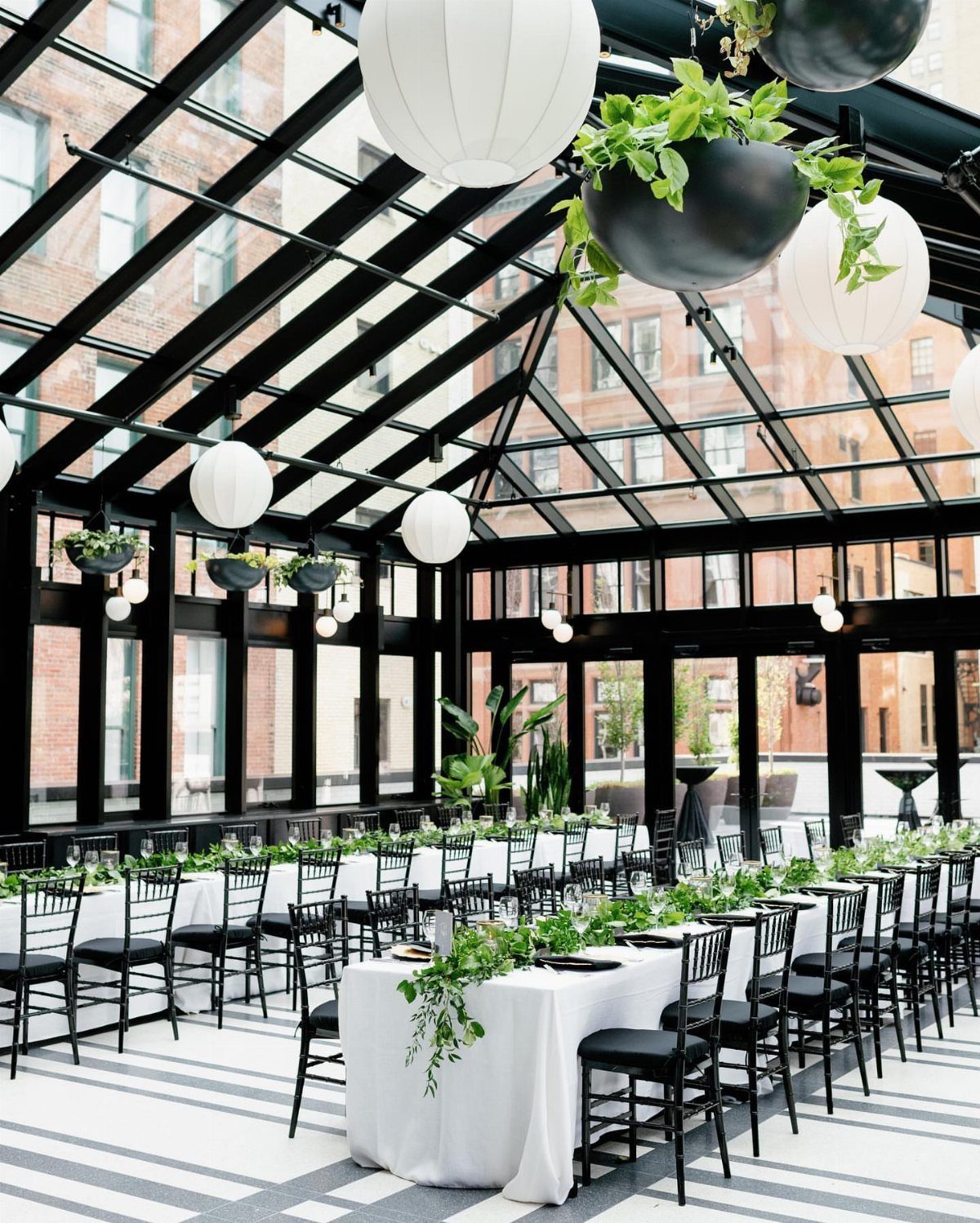Long wedding table with white linens and greenery in a glass-walled room.