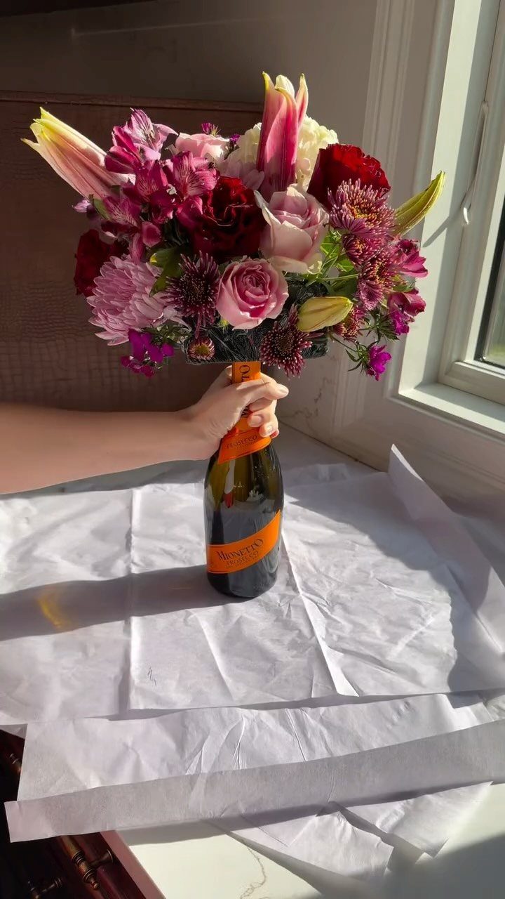 Champagne bottle topped with a lush bouquet of pink and red flowers on tissue paper.