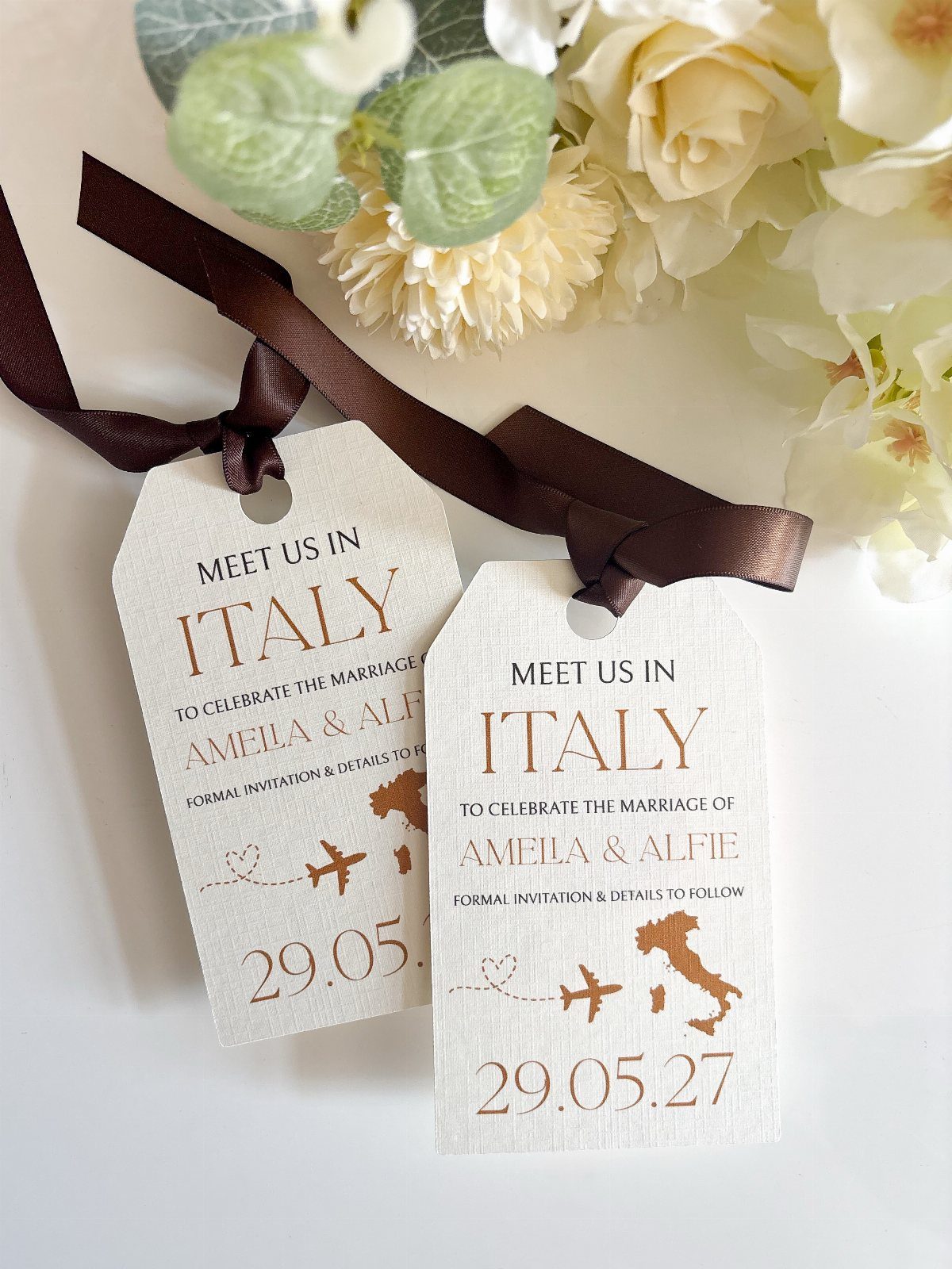 Two cream luggage tag save the dates tied with brown ribbon near white flowers.