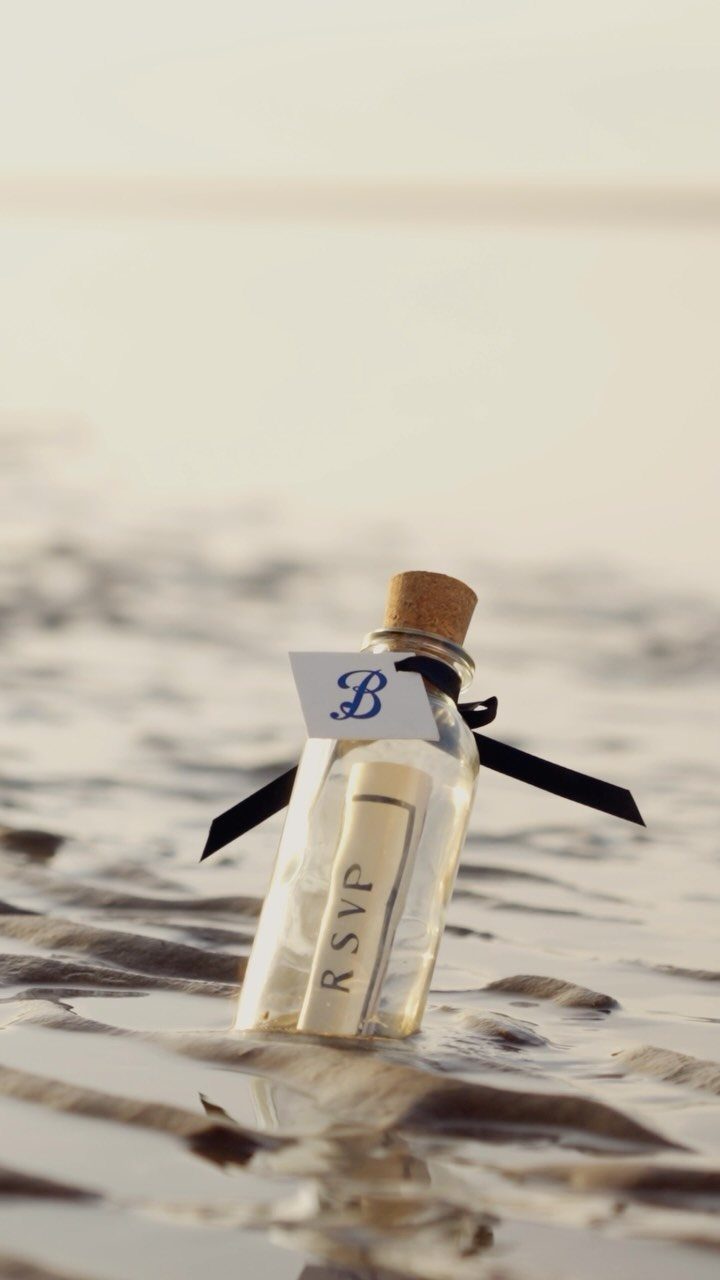 Glass bottle with a rolled RSVP scroll resting on a sandy beach shore.