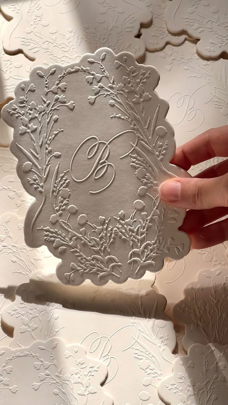 White die-cut wedding invitation with embossed floral borders and a cursive monogram.